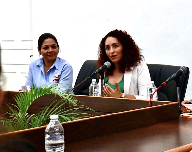 Catalina Verónica Atenco Pérez, síndica municipal de Cuernavaca, participa en una mesa de diálogo. Viste un blazer blanco y una blusa verde, mientras habla frente a un micrófono. A su lado, una representante de la CDHEM observa con atención. La escena ocurre en un recinto con mobiliario de madera y decoración vegetal.