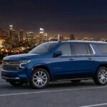 Camioneta blindada tipo Suburban color azul estacionada en mirador con vista panorámica de una ciudad iluminada de noche, unidad lista para operación de movilidad segura de Shel-Ha Soluciones.